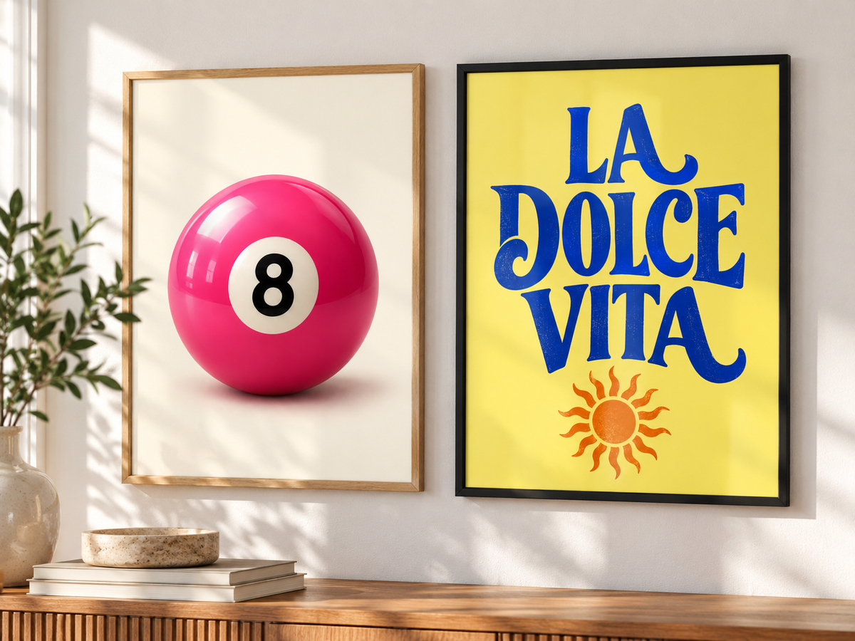 Two framed wall art prints displayed side by side on a white wall above a wooden console table. The left print has a light wood frame and shows a glossy pink pool ball with the number 8.