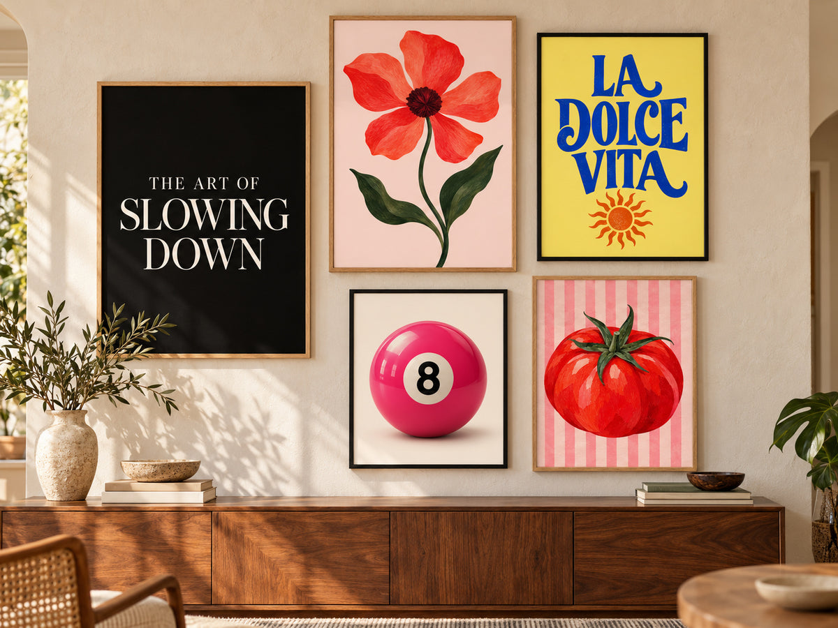 A styled interior wall with five framed art prints above a wooden console. The prints include black-and-white text reading 'THE ART OF SLOWING DOWN,' a pink flower on a blush background, blue text reading 'LA DOLCE VITA' on yellow, a pink billiard ball with the number 8, and a red tomato on pink stripes.