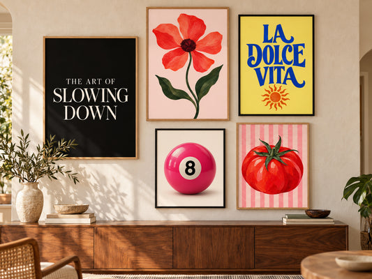 A styled interior wall with five framed art prints above a wooden console. The prints include black-and-white text reading 'THE ART OF SLOWING DOWN,' a pink flower on a blush background, blue text reading 'LA DOLCE VITA' on yellow, a pink billiard ball with the number 8, and a red tomato on pink stripes.