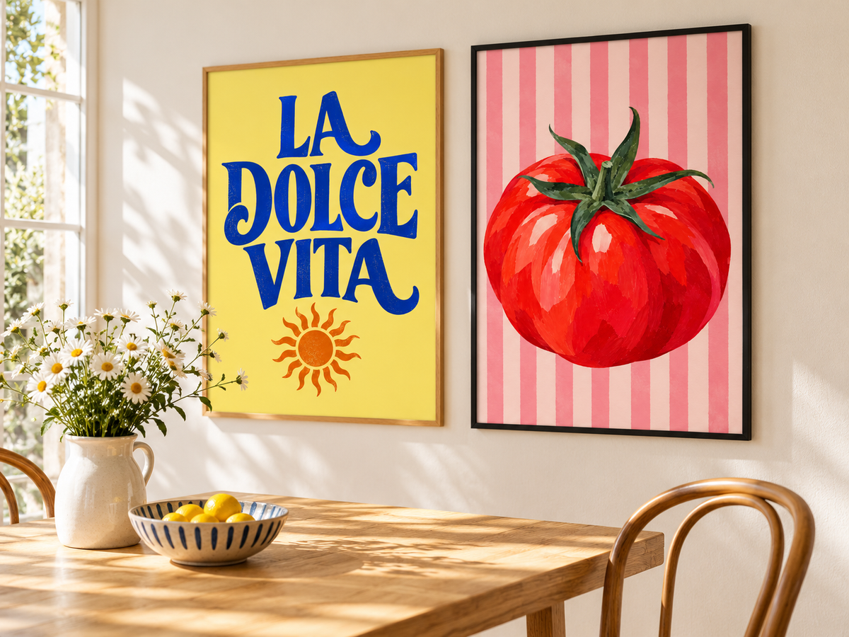 Two framed wall art prints hanging above a wooden dining table in a bright room. The left print has a yellow background with blue text reading 'LA DOLCE VITA' and an orange sun, while the right print shows a large red tomato on pink vertical stripes.