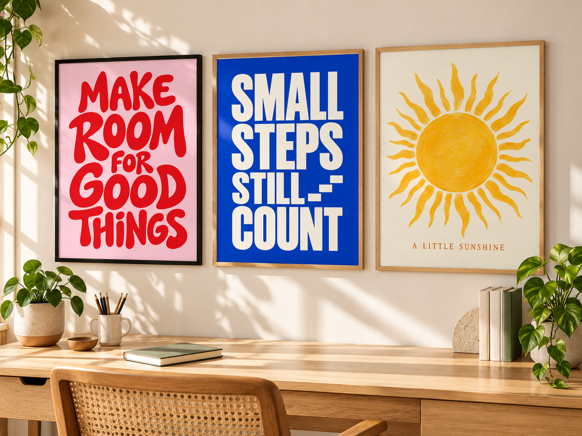 Three framed motivational wall art prints hang above a wooden desk in a bright, sunlit room. The left print has red text on a pink background reading “MAKE ROOM FOR GOOD THINGS,” the middle print has white text on a blue background reading “SMALL STEPS STILL COUNT,” and the right print shows a yellow sun on an off-white background with the text “A LITTLE SUNSHINE.