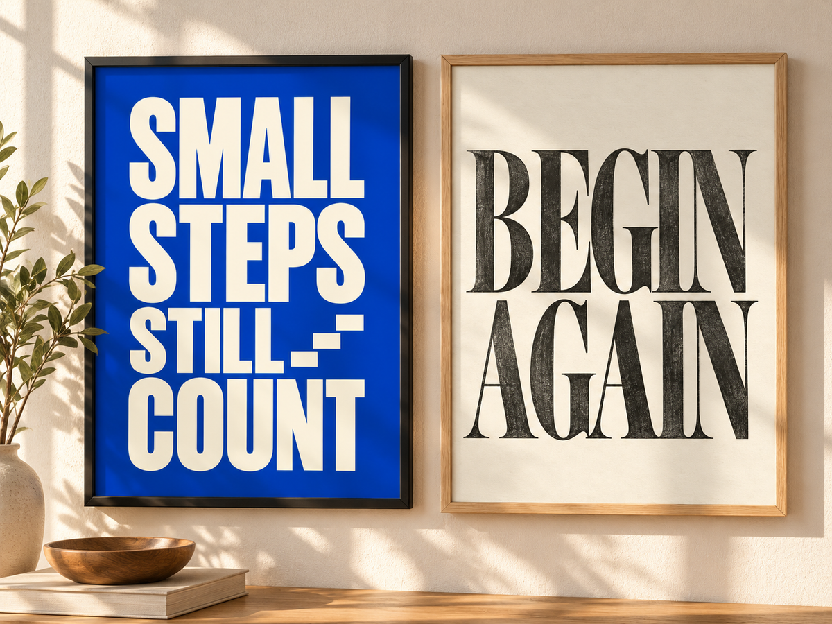 Two framed motivational wall art prints displayed side by side on a beige wall above a wooden surface. The left print has a black frame and a bright blue background with bold white text reading 'SMALL STEPS STILL COUNT.