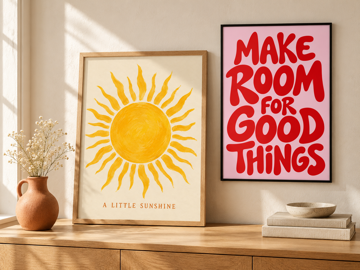 Two framed art prints displayed on a beige wall above a wooden console table. The left print features a yellow hand-drawn sun on an off-white background with the text 'A LITTLE SUNSHINE' at the bottom.