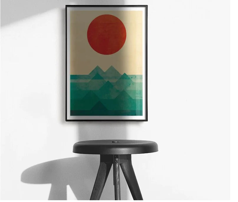Framed abstract art with a red circle and green mountains on a white wall above a black stool.