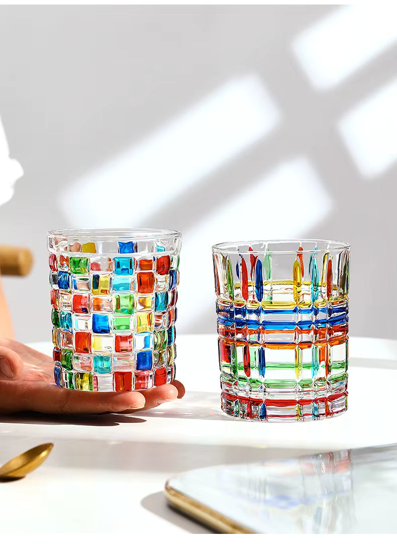 Two clear glass tumblers with colorful mosaic and striped cut-glass patterns, shown on a white tabletop with a hand holding one tumbler. The background is a bright white wall with soft window-light shadows.