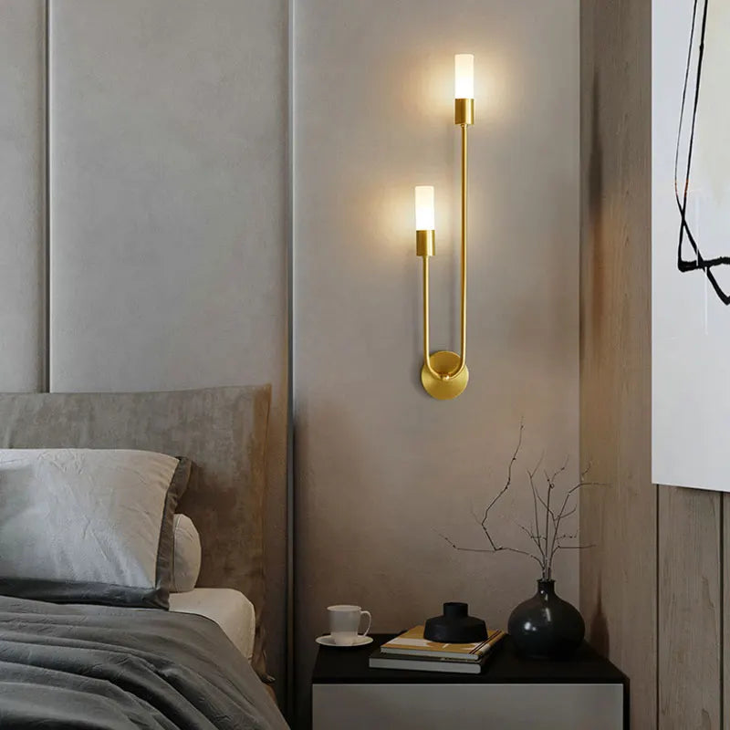 Gold wall sconce on a bedroom wall with a bed and nightstand in the foreground.