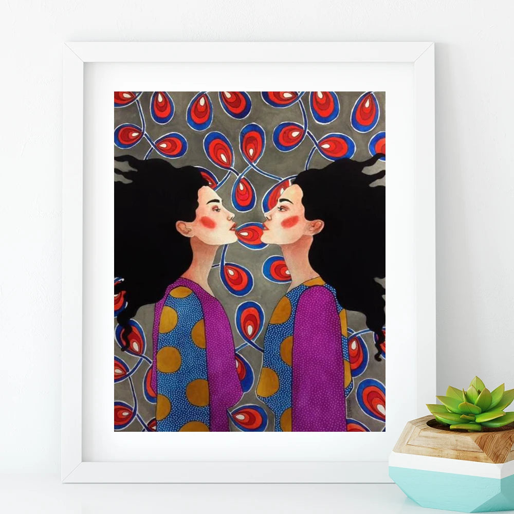 Framed artwork of two women blowing kisses against a patterned background, with a small plant on a white surface.