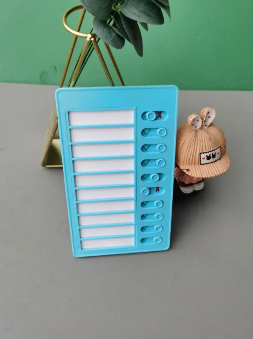 Blue reusable habit tracker board with sliding toggles, displayed on a table next to a doll and small plant in a gold stand