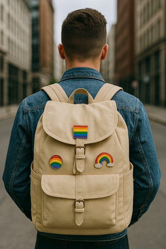 Rear view of a person in a denim jacket carrying a beige canvas backpack adorned with three rainbow pride pins, standing on a blurred urban street.