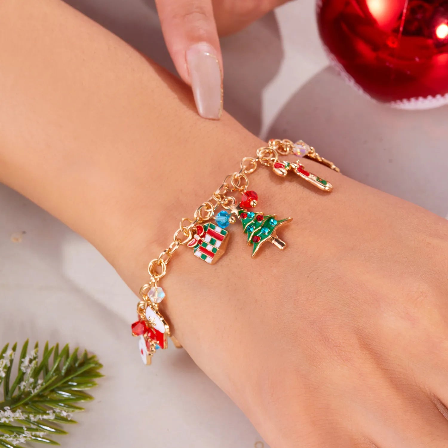Gold bracelet with Christmas-themed charms on a wrist, with a blurred red ornament and greenery in the background.