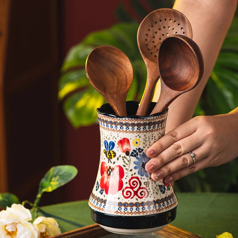 Decorative ceramic utensil holder with wooden spoons held by a hand, against a blurred green plant background.
