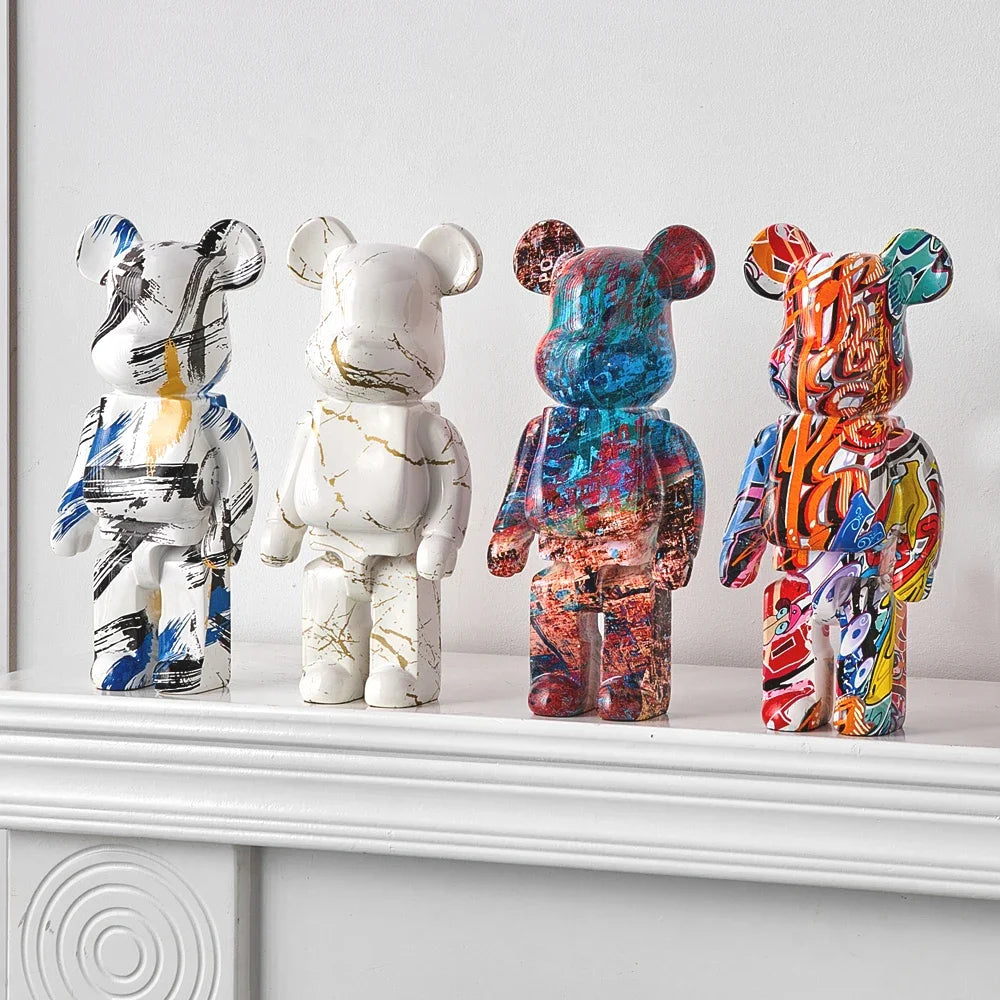 Four bear-shaped collectible figurines displayed side by side on a white mantelpiece against a light gray wall. The leftmost figure has a white base with blue, black, and gold abstract paint strokes; the second is white with gold marble veining; the third is multicolored with blue, red, and orange tones; the rightmost is highly colorful with orange, blue, yellow, and red abstract patterns.