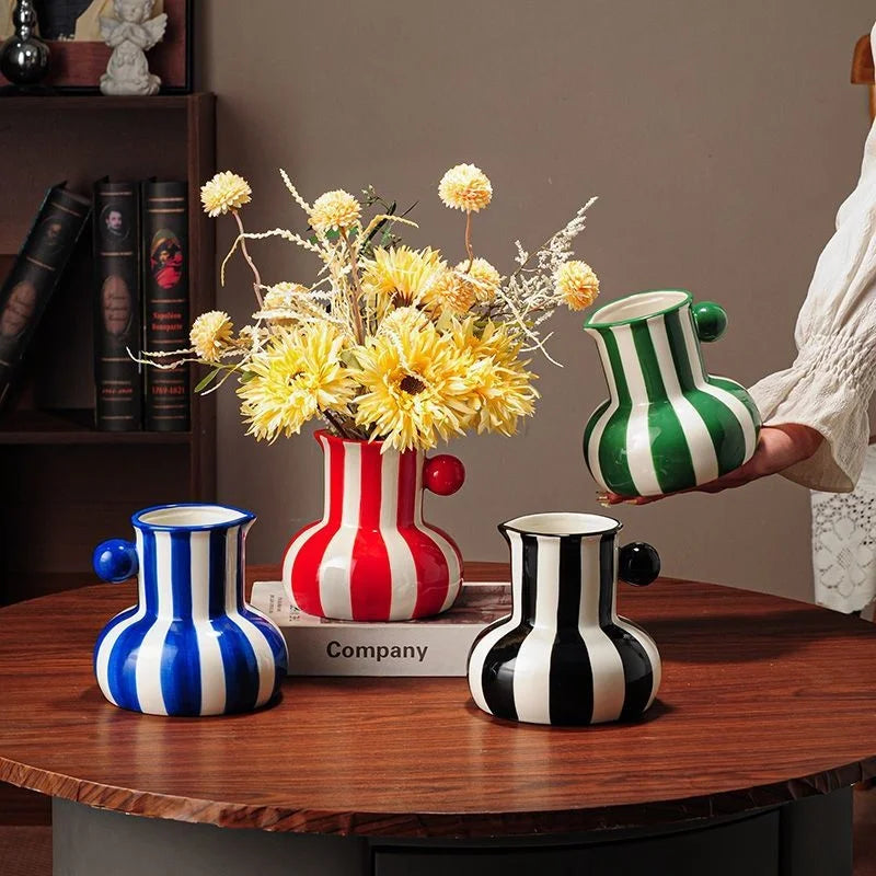 Three ceramic pitchers with bold vertical stripes in blue, red, and green-and-white, arranged on a wooden table with yellow flowers in the center. A person in a white blouse holds the green-and-white pitcher on the right.