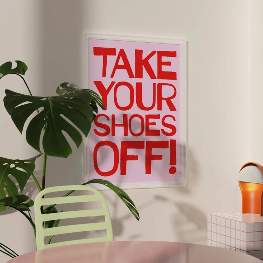 Framed poster with 'Take Your Shoes Off!' text on a wall next to a plant and chair.