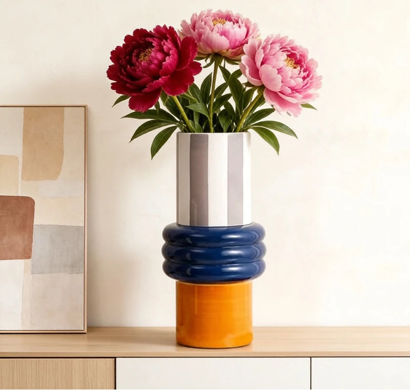 A modern stacked ceramic vase with a silver top, dark blue ribbed middle section, and orange base, holding pink and burgundy peony flowers on a light wood surface against a cream wall.