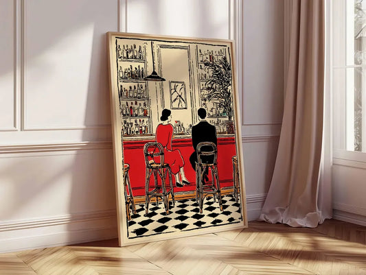 A framed art print leaning against a white paneled wall on a light wood floor, depicting a vintage bar scene with two people seated at a red counter and a black-and-white checkered floor.