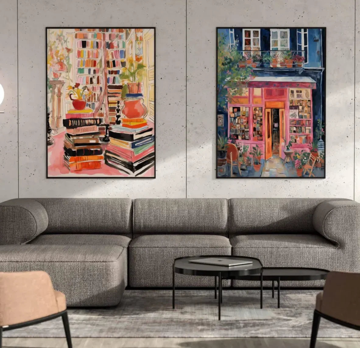 Two framed colorful paintings displayed side-by-side on a light gray concrete wall above a modern gray sectional sofa. The left painting depicts a stack of books and a red vase, while the right painting shows a vibrant pink storefront with a street scene.