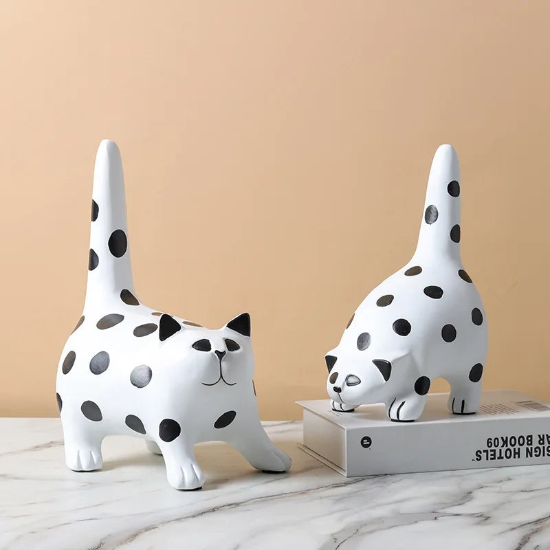 Two white cat-shaped figurines with black spots on a marble surface against a beige wall.