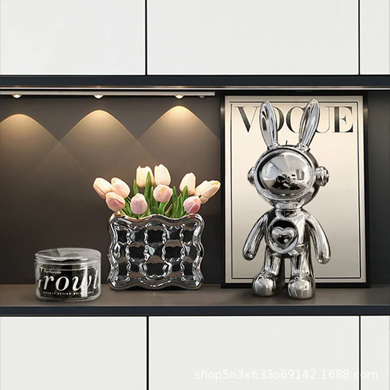 Decorative shelf with a glass vase, candle, and metallic rabbit sculpture against a dark wall with a framed magazine.