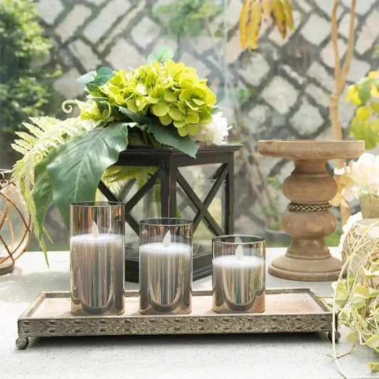 Decorative setup with candles in glass holders on a metal tray, surrounded by greenery.