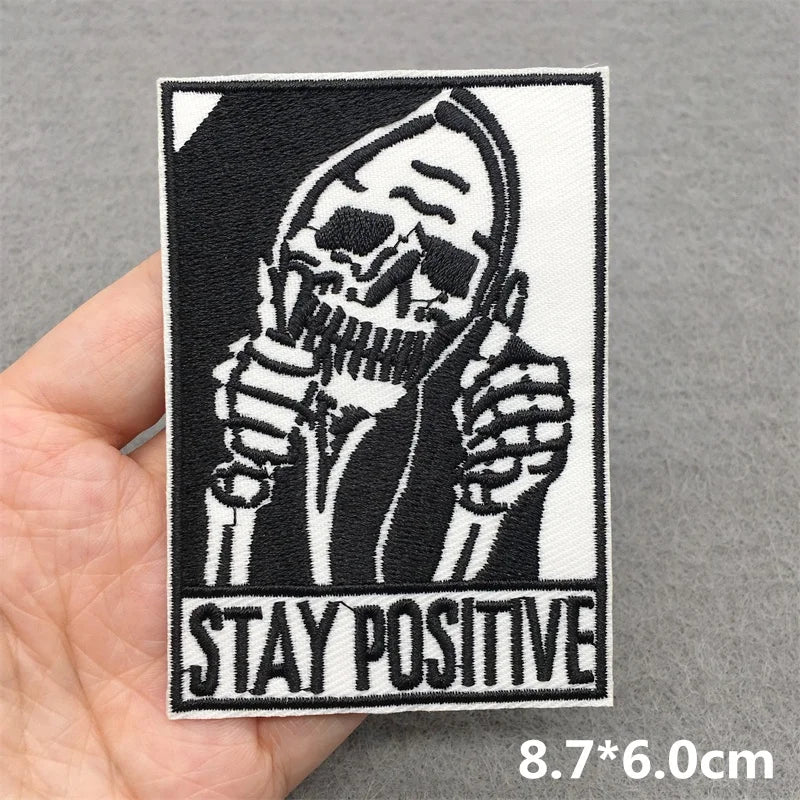 Patch with skeleton holding a microphone and 'Stay Positive' text, held by a hand on a gray background.