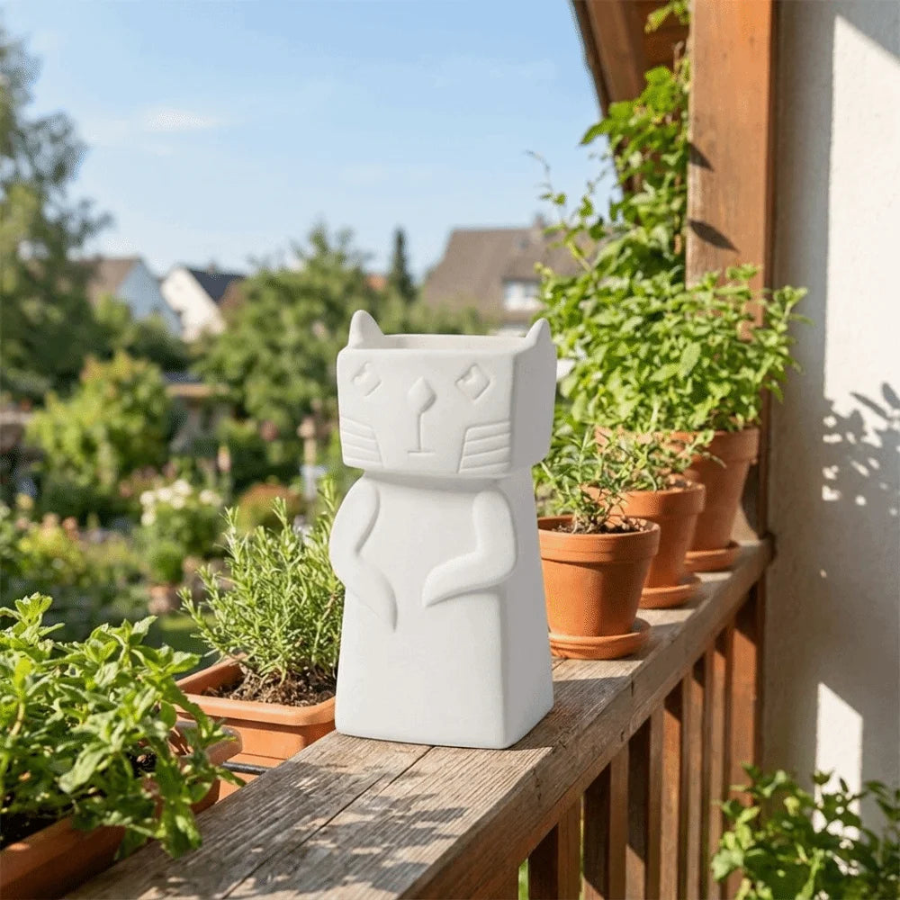 A white ceramic cat-shaped garden light or planter sits on a wooden balcony railing surrounded by potted green plants, with a blurred outdoor garden background.