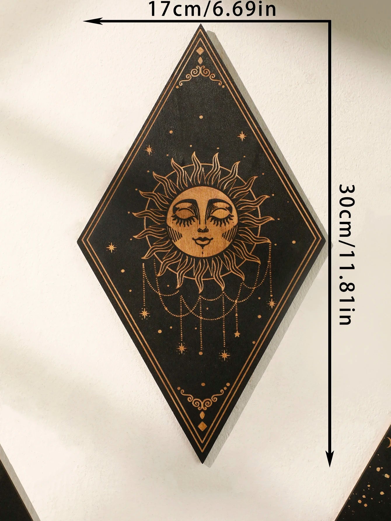 A diamond-shaped black wall art piece featuring a gold sun face with closed eyes, surrounded by stars and hanging chains, shown with dimension arrows and text indicating 17cm by 30cm.