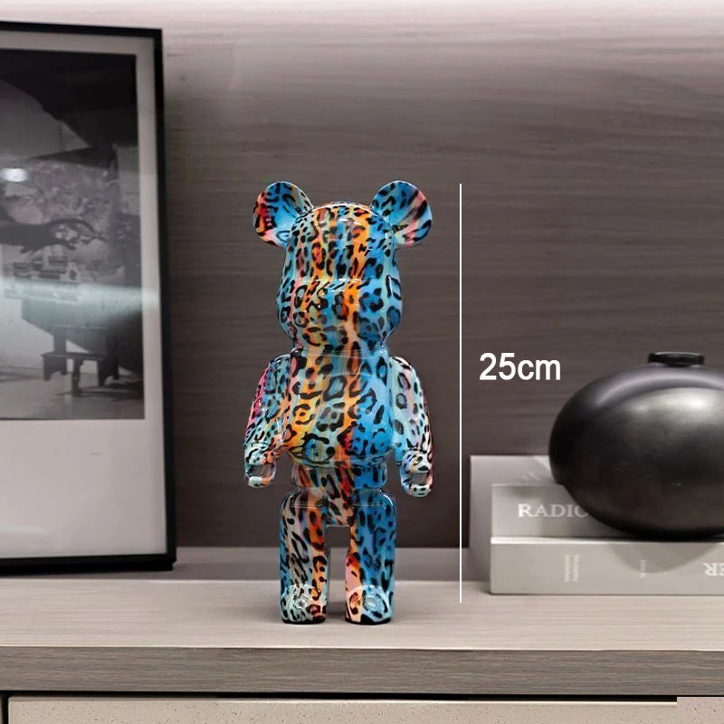 A colorful bear-shaped collectible figure with a blue, orange, and black leopard-style pattern stands on a shelf in a styled interior setting. A vertical measurement marker reads 25cm next to the figure.