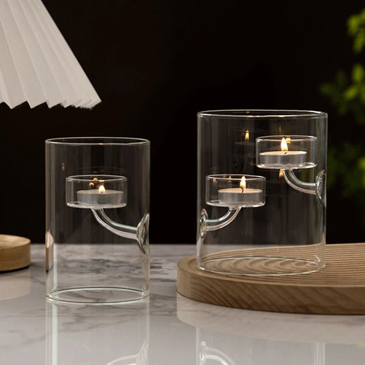 Two clear glass candle holders with candles on a reflective surface.