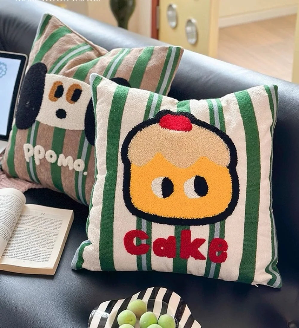 Two decorative throw pillows on a black leather sofa. The front pillow has a green and white striped pattern with a yellow cupcake character and the word 'Cake' in red.
