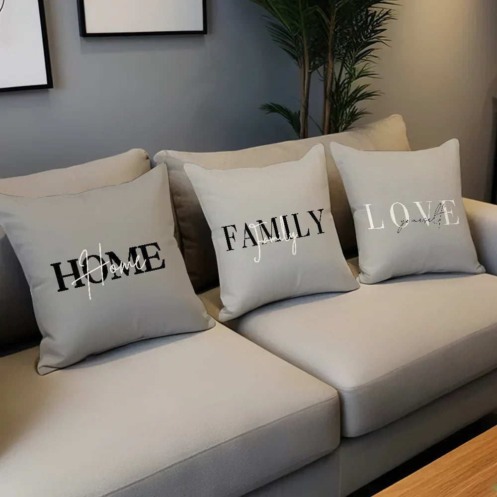Three light gray throw pillows with black and white text reading HOME, FAMILY, and LOVE, arranged on a light beige sofa in a living room setting.