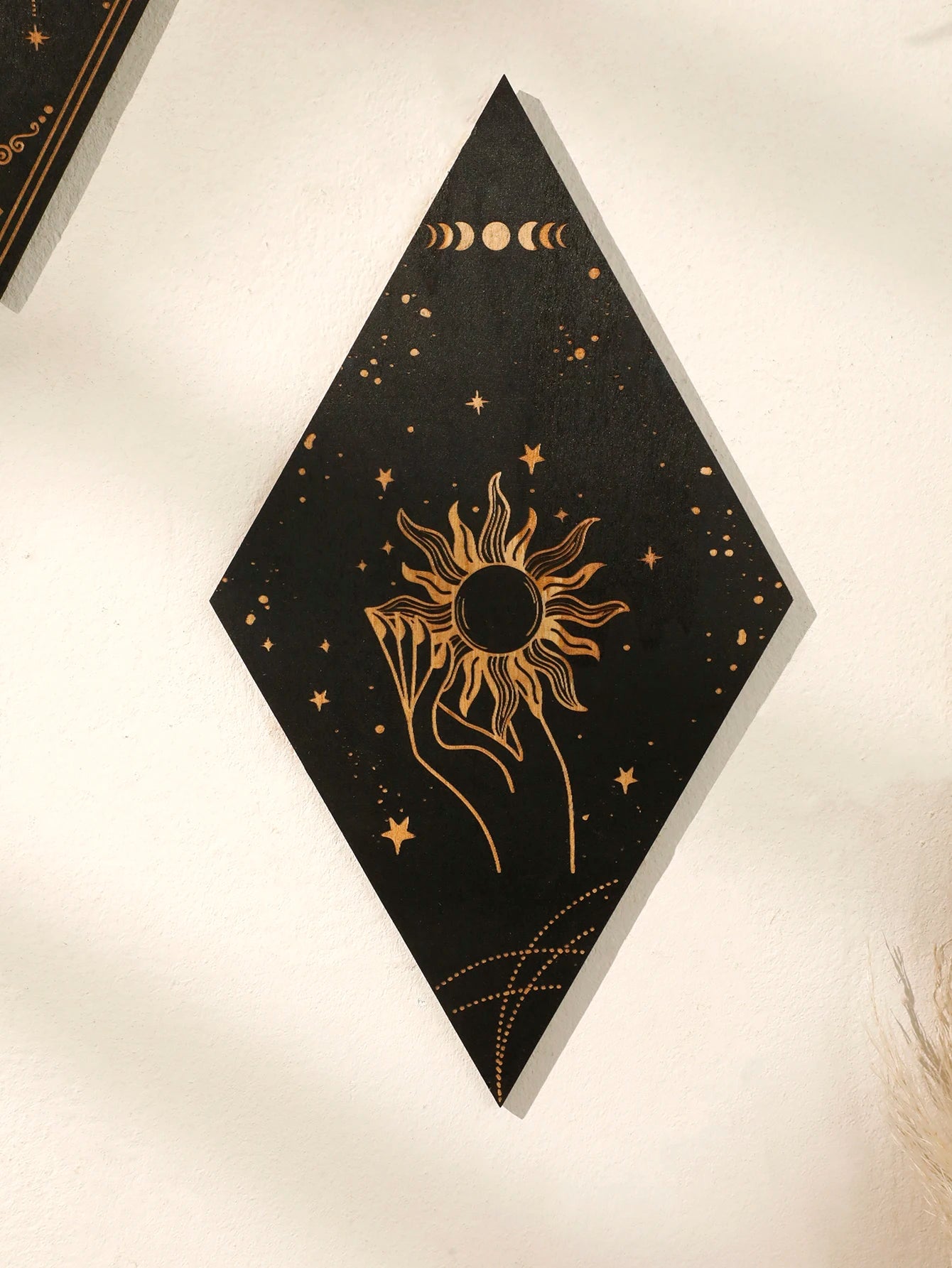 A black diamond-shaped wall art piece with gold celestial designs, including a sun, moon phases, stars, and a hand, displayed on a textured off-white wall.