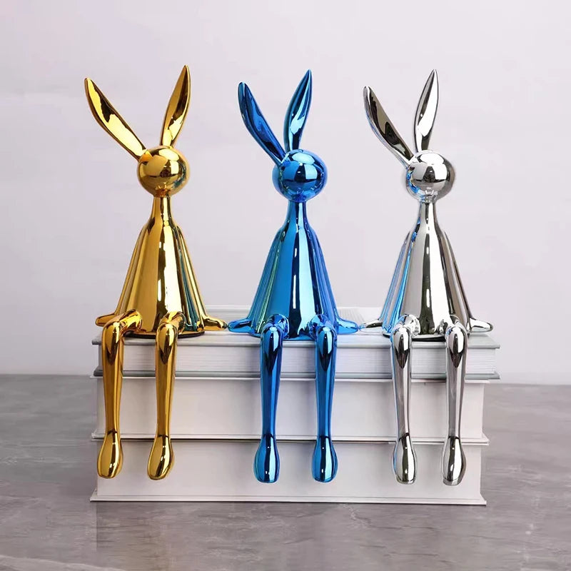 Gold, blue and silver rabbit ornaments sitting on stacked white books, with tall ears and long legs dangling over the edge.