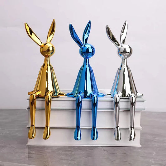 Gold, blue and silver rabbit ornaments sitting on stacked white books, with tall ears and long legs dangling over the edge.