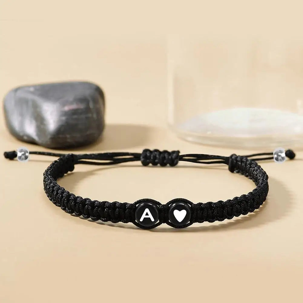 Black braided bracelet with letter 'A' and heart charm on a beige surface.