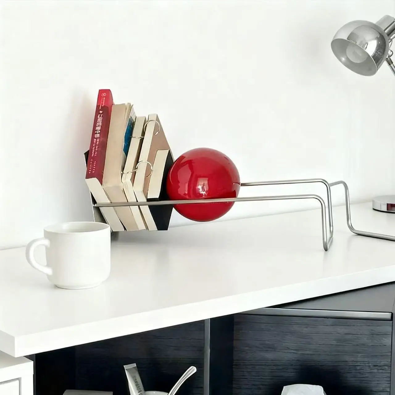 Modern metal desktop bookshelf with red decorative sphere holding books on a contemporary white desk.