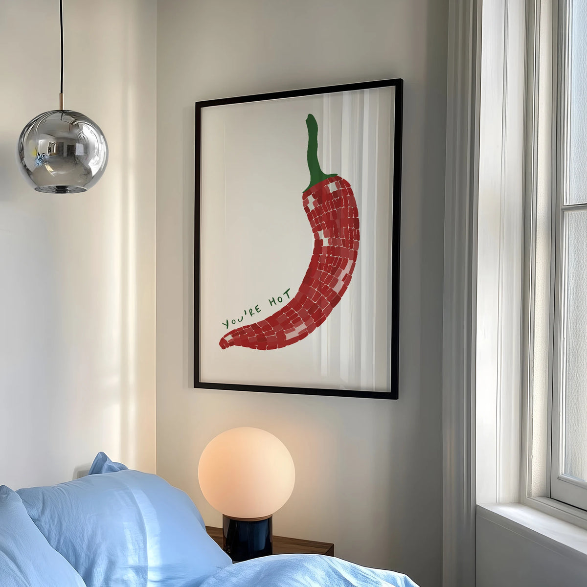 Framed artwork of a red chili pepper on a wall above a bed with a lamp.
