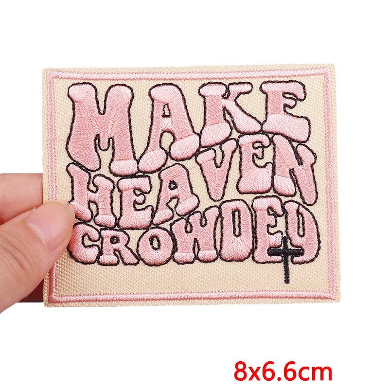 Embroidered patch with 'MAKE HEAVEN CROWDED' text held by a hand, on a beige background.
