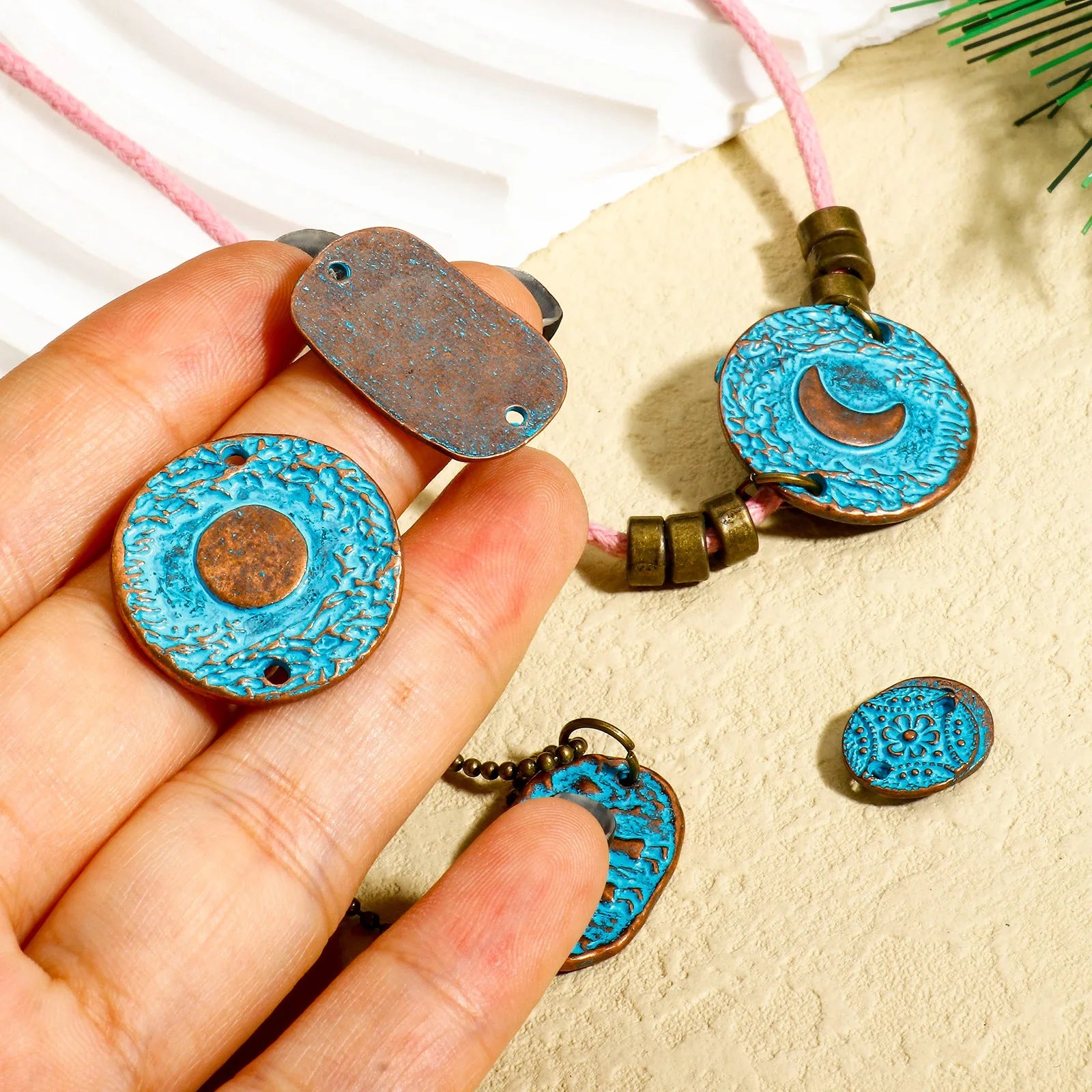 Hand holding a blue and bronze decorative pendant with more pendants on a beige surface.