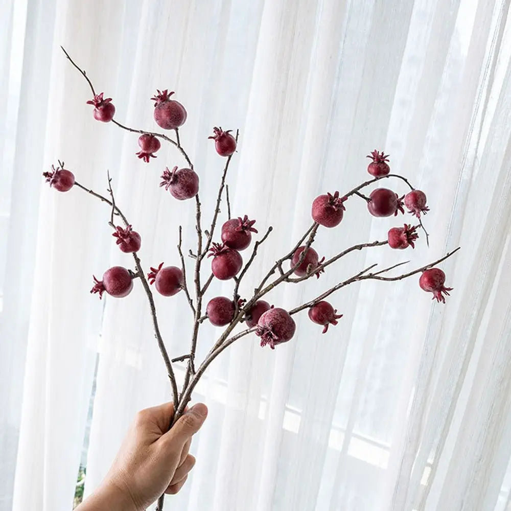 Hand holding a decorative branch with red fruit-like pods against a white curtain background