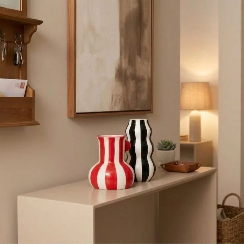 Blue and white rounded vase with beaded handles, tall black and white wavy vase holding white flowers, and red and white rounded vase on a wooden sideboard.