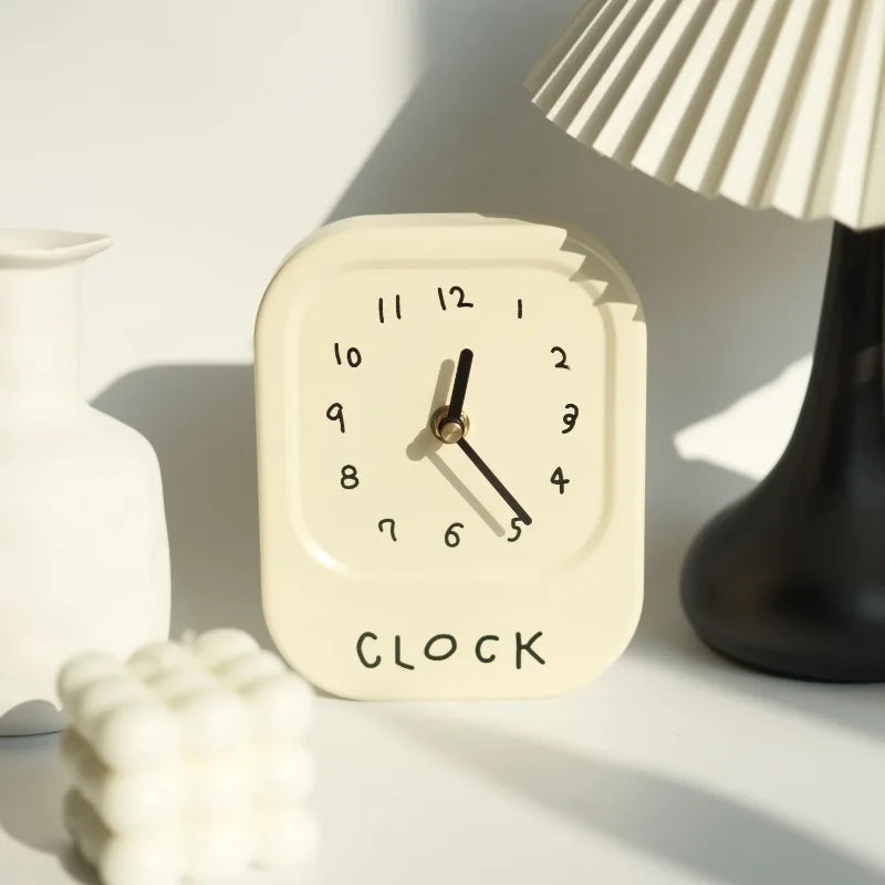 Small beige clock with 'CLOCK' text on a white surface with a lamp in the background