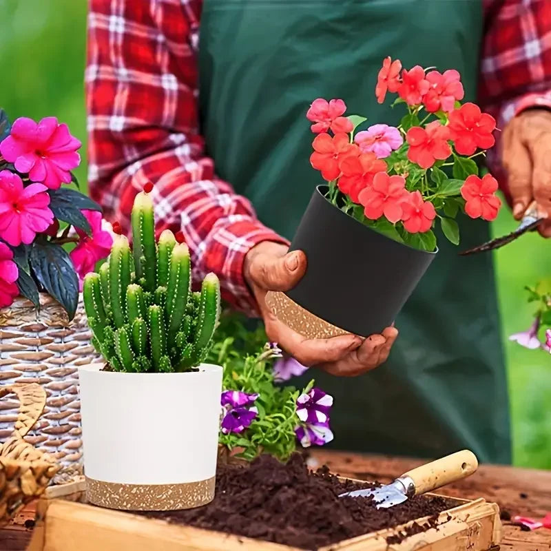 Person holding a potted plant with pink flowers, surrounded by other plants and gardening tools.