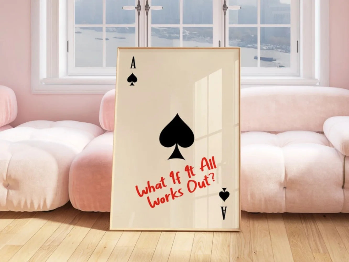 Framed artwork with playing card symbols and text 'What If It All Works Out?' in a room with pink couches.