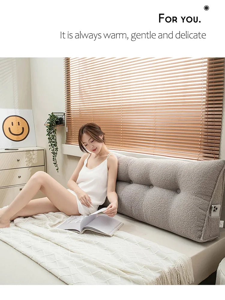 Woman reading a book on a bed with a large gray pillow, smiling emoji on a shelf in the background.