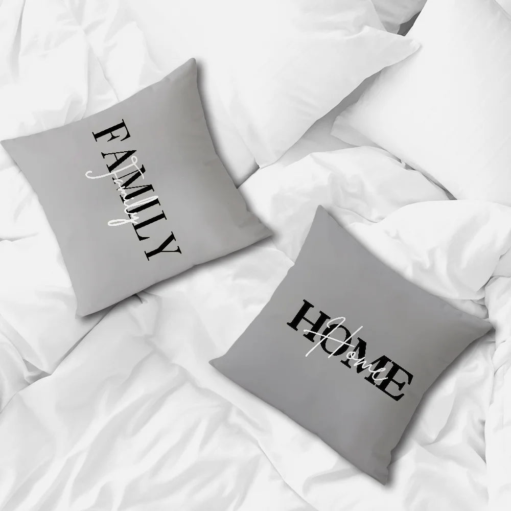 Two square gray throw pillows on a white bed. One pillow reads 'FAMILY' with a script overlay, and the other reads 'HOME' with a script overlay.