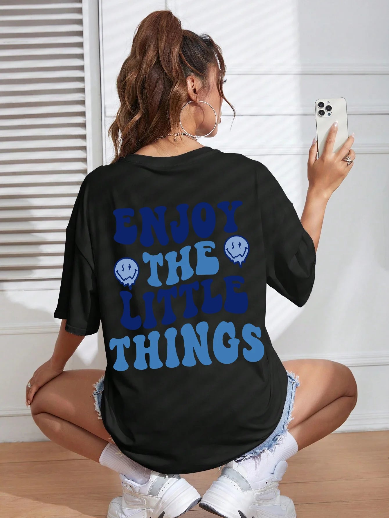 Person wearing a black t-shirt with 'Enjoy the Little Things' text and smiley faces, sitting on the floor.