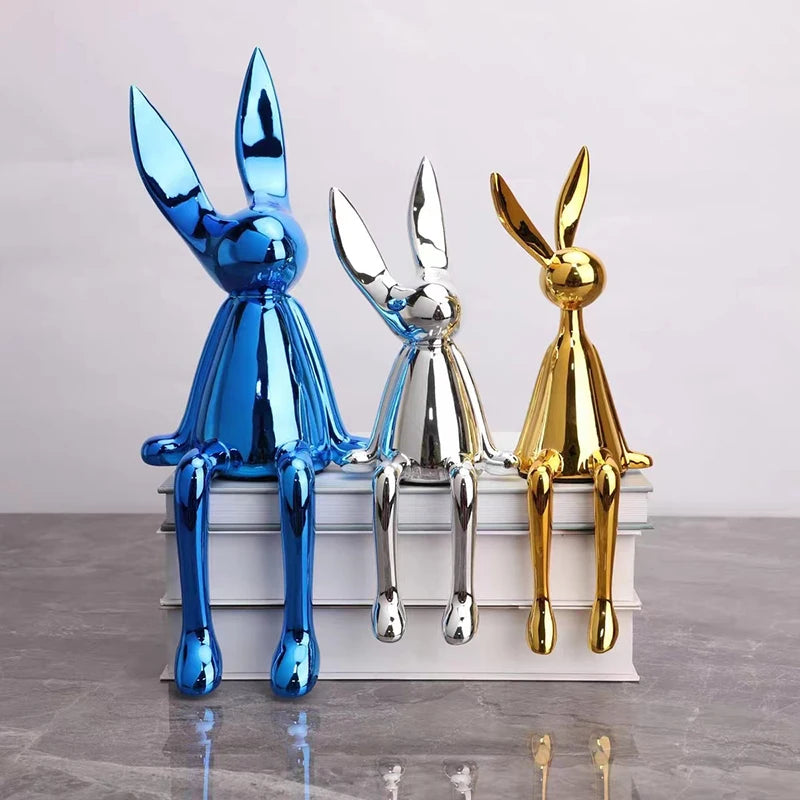Three metallic rabbit sculptures in blue, silver, and gold on a white pedestal against a light gray background.