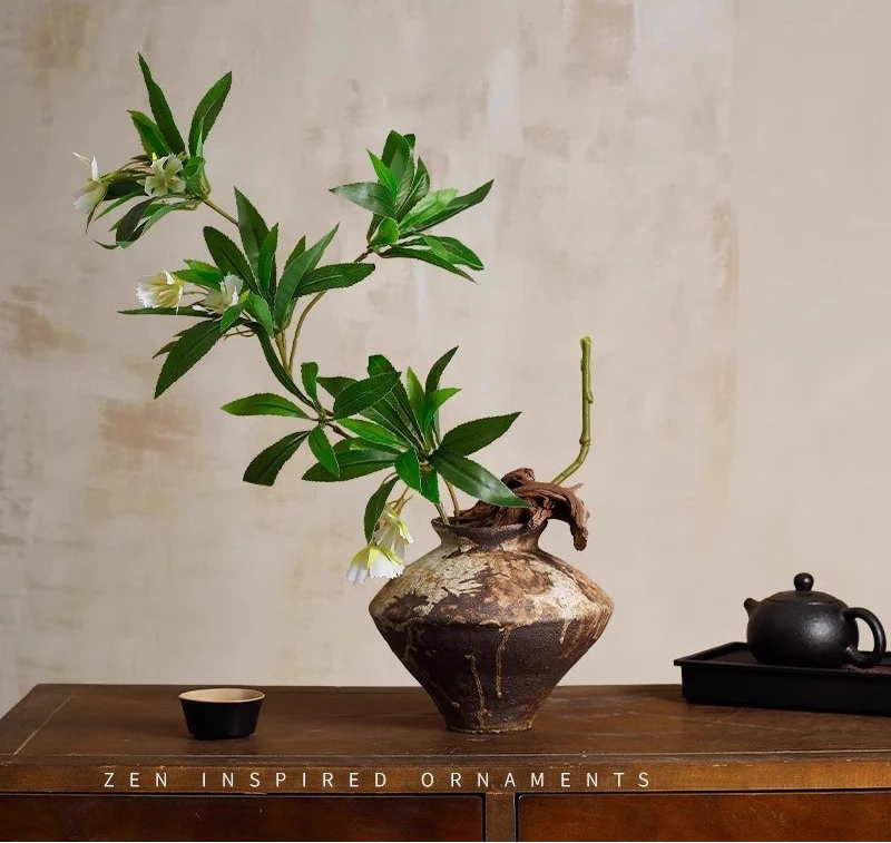 A decorative artificial plant with green leaves and small white flowers arranged in a rustic brown Wabi-sabi ceramic vase on a wooden table, with a black teapot on a tray in the background.