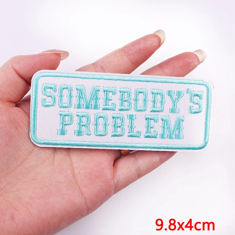Rectangular patch with 'SOMEBODY'S PROBLEM' text held in a hand on a white background